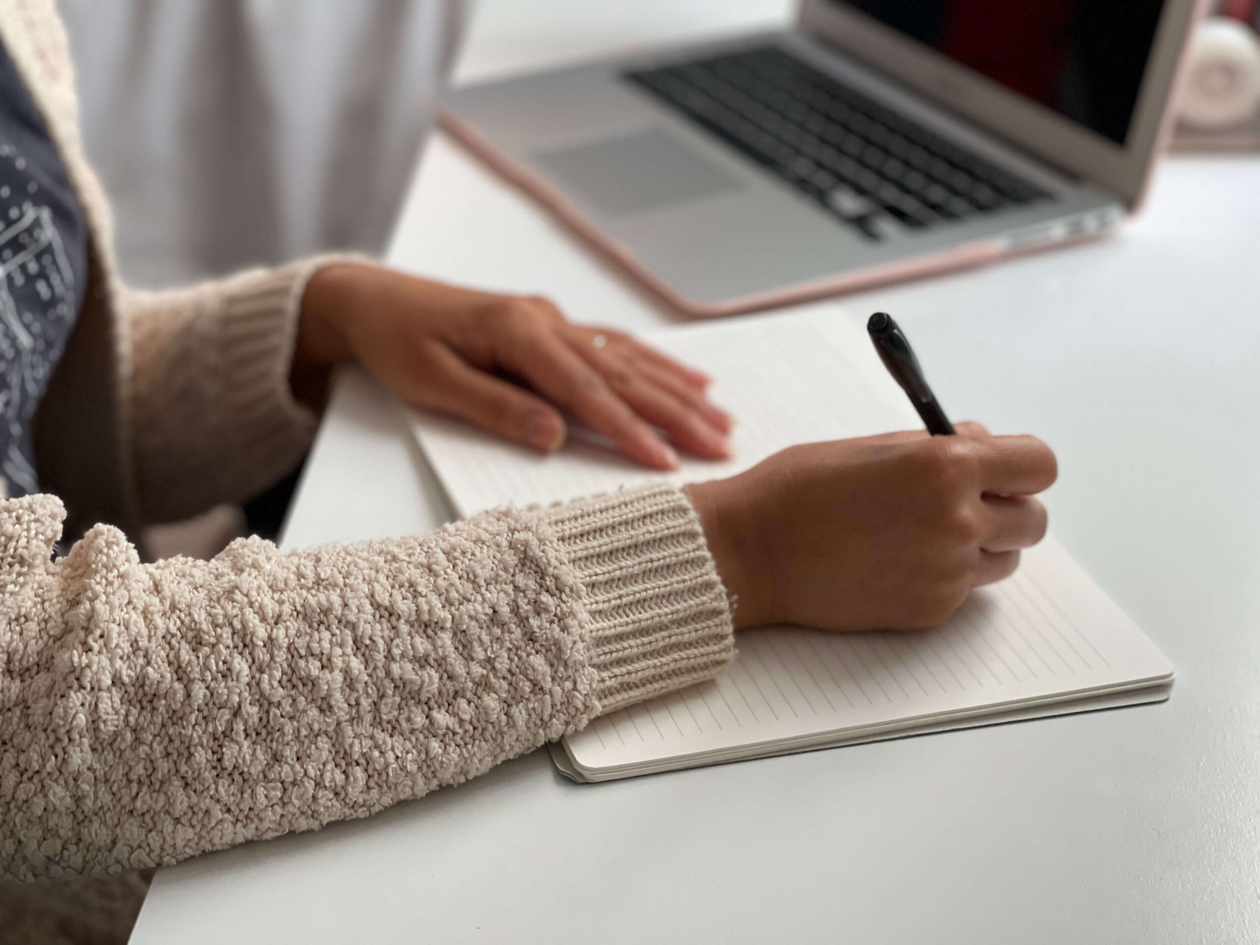 Woman wearing gray shirt and cream cardigan journaling on a white desk.