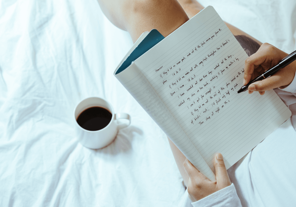 Woman writing in a teal colored journal on her bed with a pen in her hand and coffee sitting next to her.